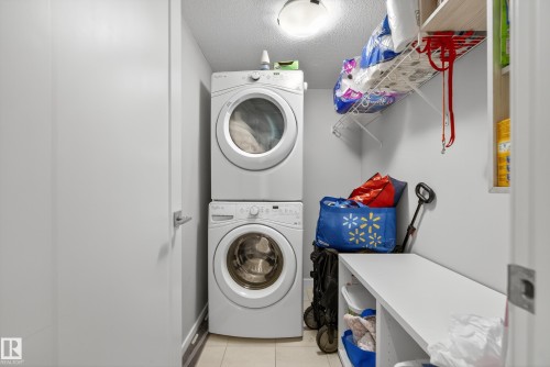 Dedicated laundry space featuring a stacked washer and dryer, ceiling-mounted light fixture, built-in shelving, and tile flooring - #107 1031 173 St Sw, Edmonton, AB 