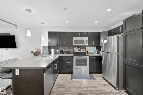 Contemporary kitchen featuring dark cabinetry, stainless steel appliances, light-toned countertops, and wood-finish flooring - #107 1031 173 St Sw, Edmonton, AB 