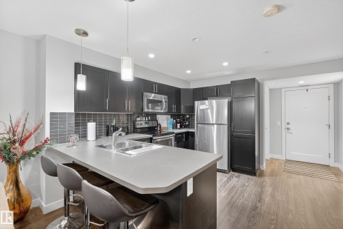Kitchen featuring dark cabinetry, stainless steel appliances, a tiled backsplash, and an island with an integrated sink - #107 1031 173 St Sw, Edmonton, AB 
