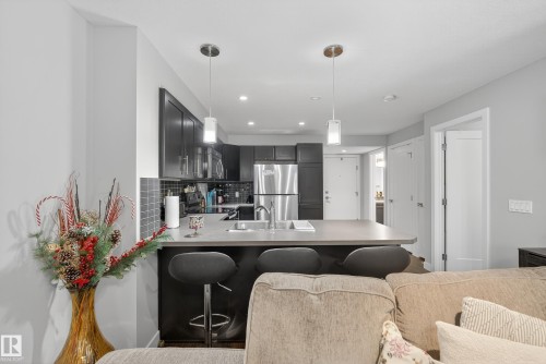 Open-concept kitchen featuring dark wood cabinetry, stainless steel appliances, a grey tile backsplash, and a breakfast bar with pendant lighting - #107 1031 173 St Sw, Edmonton, AB 