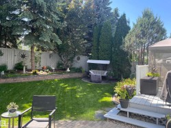 Expansive backyard featuring a lush lawn, mature trees, a raised garden bed with retaining wall, a paved patio area, and a wood deck - 