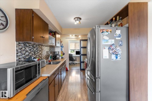 Galley kitchen featuring wood-finish flooring, dark wood upper and lower cabinetry, a mosaic tile backsplash, and a stainless steel refrigerator - 173 Cornell Court Nw, Edmonton, AB - Indoor Photo Showing Kitchen