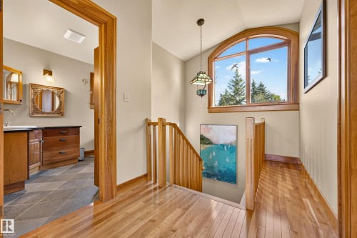 Hardwood flooring throughout, arched window with natural wood trim, wood-finish staircase railing, pendant light fixture, and an adjacent bathroom with a wood vanity and stone-look tiled flooring - 51 Itaska Beach, Rural Leduc County, AB 
