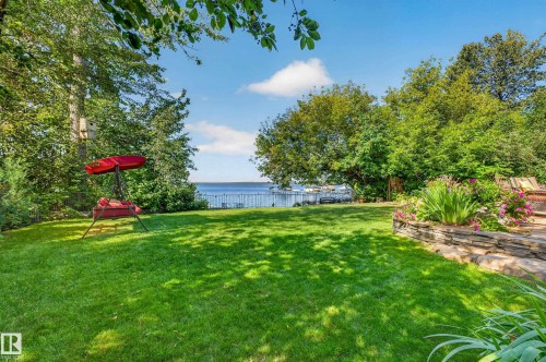 Expansive lawn area, mature trees, stone retaining wall with plantings, water body in the background, perimeter fencing - 51 Itaska Beach, Rural Leduc County, AB 