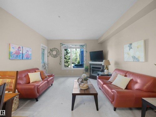 Carpeted living space featuring a corner fireplace with a tiled surround, a large window, and light-toned walls - #206 12111 51 Av Nw, Edmonton, AB 