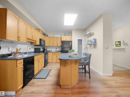 Kitchen featuring wood-finish cabinetry, dark countertops, black appliances, a central island with seating, and wood-finish flooring - #206 12111 51 Av Nw, Edmonton, AB 