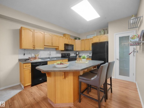 Kitchen featuring wood-finish flooring, light wood cabinetry, black appliances, a central island with seating, and a recessed ceiling light - #206 12111 51 Av Nw, Edmonton, AB 