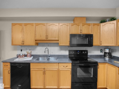 Kitchen featuring wood cabinetry, a dual-basin stainless steel sink, black appliances, solid surface countertops, and a tiled backsplash - #206 12111 51 Av Nw, Edmonton, AB 