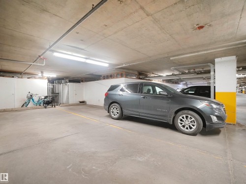 Concrete-finish parking surface, fluorescent overhead lighting, exposed ceiling pipes, yellow accent pillar, white storage walls - #206 12111 51 Av Nw, Edmonton, AB 