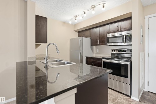 Kitchen featuring a double basin stainless steel sink, stainless steel appliances, dark wood-finish cabinetry, and track lighting - #225 1510 Watt Dr Sw, Edmonton, AB 