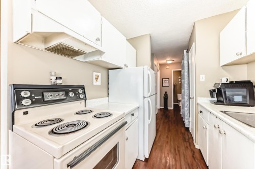 Galley kitchen featuring a white electric range, white upper and lower cabinetry, white refrigerator, and wood-finish flooring - #302 10606 123 St Nw, Edmonton, AB 