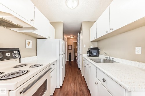 Galley kitchen featuring wood-finish flooring, white cabinetry, integrated range, refrigerator, and stainless steel sink - #302 10606 123 St Nw, Edmonton, AB 