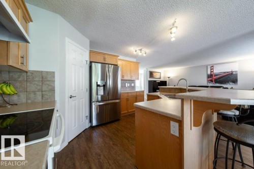 Kitchen featuring wood-finish cabinetry, stainless steel refrigerator, tile backsplash, and an island with bar seating - 16120 138 Street Nw, Edmonton, AB 