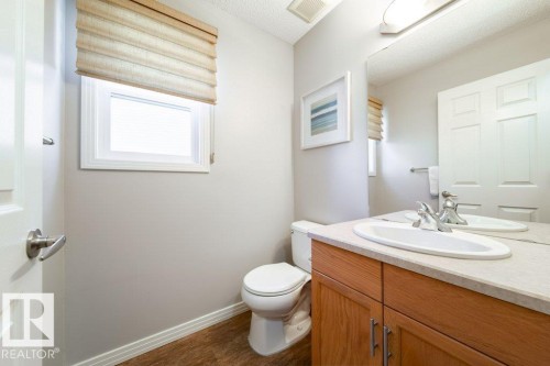 Bathroom featuring a wood-finish vanity with an integrated sink, a wall-mounted mirror, a window with a Roman shade, and wood-finish flooring - 16120 138 Street Nw, Edmonton, AB 