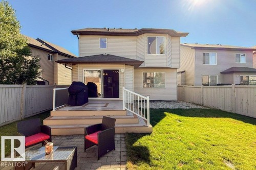 Rear exterior featuring light-toned siding, a multi-level deck with glass panel railings, a ground-level paver patio, a sliding glass door, and a fenced yard with established lawn - 16120 138 Street Nw, Edmonton, AB 