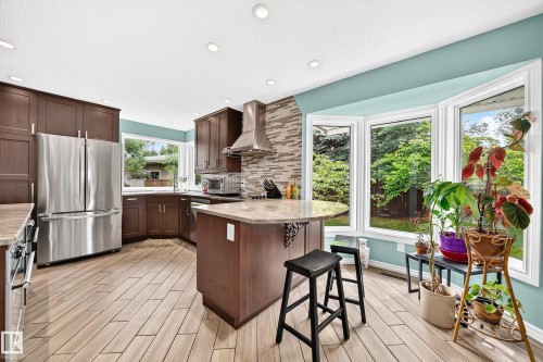 Kitchen featuring wood-finish flooring, dark wood cabinetry, stainless steel appliances, a stone countertop island, and a bay window - 8224 186 Street Nw, Edmonton, AB 
