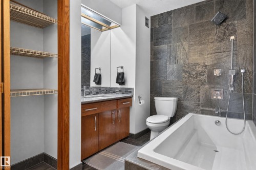 Full bathroom featuring a single vanity with wood cabinetry, a built-in tub with a shower fixture, and dark stone tile walls - 17103 92 Street Nw, Edmonton, AB 