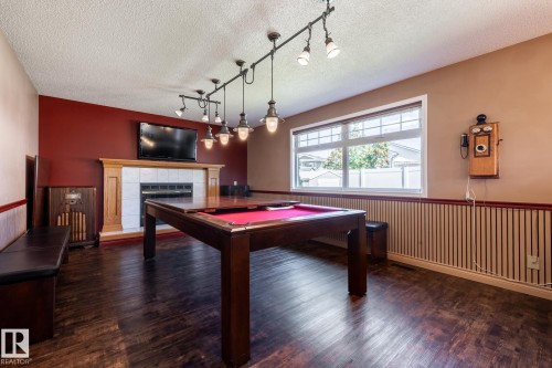Wood-finish flooring, a fireplace with a white tile surround and wood mantel, a large window with white blinds, and track lighting with multiple pendant fixtures - 89 Ozerna Road Nw, Edmonton, AB 