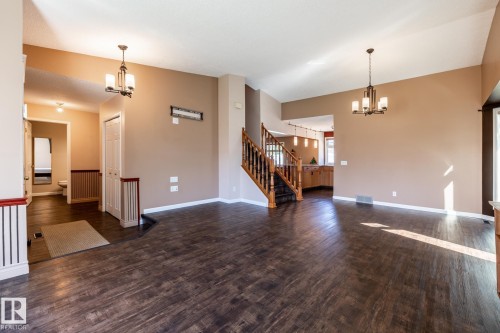 Expansive living area with wood-finish flooring, two decorative chandeliers, a natural wood staircase with wrought iron spindles, and neutral paint tones - 89 Ozerna Road Nw, Edmonton, AB 