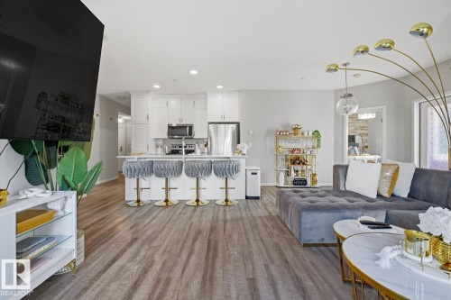 Open-concept living space featuring wood-finish flooring, a kitchen island with seating, white cabinetry, and stainless steel appliances - #155 1818 Rutherford Rd Sw, Edmonton, AB 