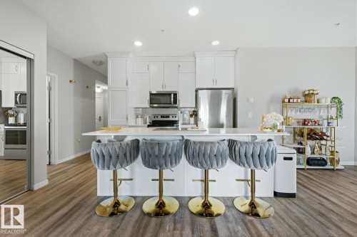 Open-concept kitchen featuring an island with a breakfast bar, white cabinetry, stainless steel appliances, and wood-finish flooring - #155 1818 Rutherford Rd Sw, Edmonton, AB 