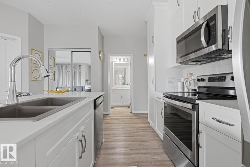 Modern kitchen featuring white cabinetry, stainless steel appliances, wood-finish flooring, a double basin sink, and white countertops - #155 1818 Rutherford Rd Sw, Edmonton, AB 
