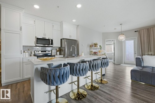 Kitchen island with white countertop, integrated stainless steel appliances, white cabinetry, and wood-finish flooring - #155 1818 Rutherford Rd Sw, Edmonton, AB 