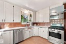 White paneled cabinetry with dark hardware, stainless steel appliances, corner sink with window, copper-toned decorative tile backsplash, and wood-finish flooring - 10943 72 Avenue Nw, Edmonton, AB 