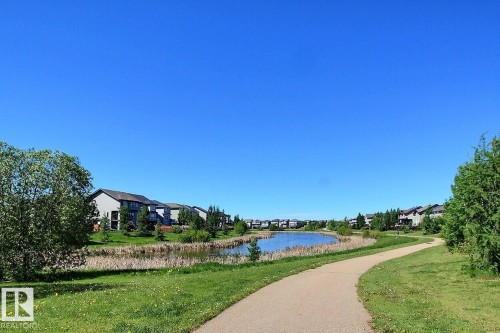 Paved pathway winding alongside a tranquil pond, bordered by green grass and mature trees - #99 1010 Rabbit Hill Rd Sw, Edmonton, AB 