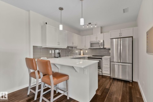 White cabinetry, stainless steel appliances, a kitchen island with seating, pendant lighting, and wood-finish flooring - #404 2755 109 St Nw, Edmonton, AB 