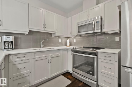 White shaker cabinetry with curved hardware, stainless steel appliances, light-toned countertops, and a gray tile backsplash - #404 2755 109 St Nw, Edmonton, AB 