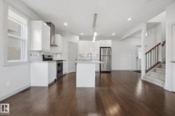 Hardwood flooring, white kitchen cabinetry, stainless steel appliances, central island with sink, and carpeted stairs with wood railing - 