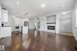 Open-concept living area featuring wood-finish flooring, a contemporary kitchen with a central island, a built-in fireplace with a stone surround, and an integrated appliance suite - 