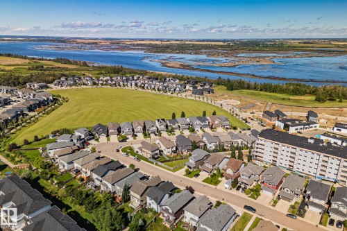 Aerial perspective of a residential community featuring a central green space, diverse housing styles, a multi-story building, and a prominent water body in the background - 2096 Redtail Common Nw, Edmonton, AB 