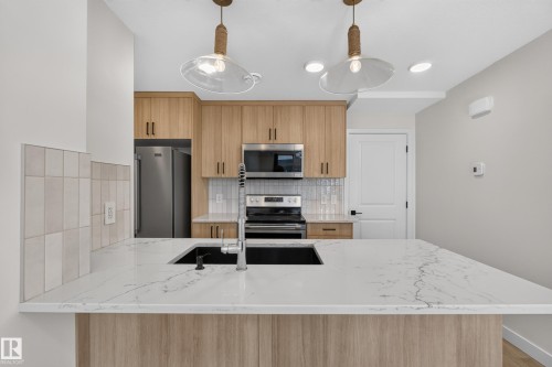 Kitchen featuring a large island with white stone-finish countertop, undermount sink, stainless steel appliances, light wood-finish cabinetry, and pendant lighting - #58 16231 19 Av Sw, Edmonton, AB 