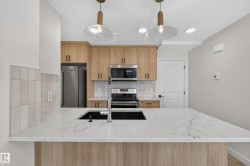 Kitchen featuring a large island with white stone-finish countertop, undermount sink, stainless steel appliances, light wood-finish cabinetry, and pendant lighting - 