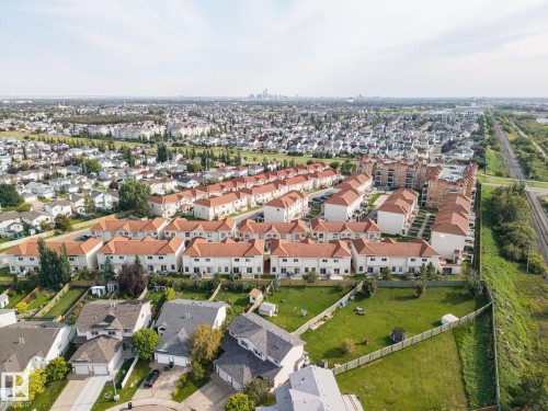 Aerial perspective showcasing a residential community with red-tiled roofs, a large grass area, and a distant city skyline - #102 13825 155 Av Nw, Edmonton, AB 