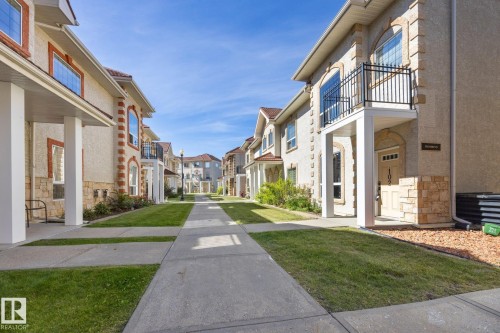 Stucco exterior with stone accents, arched windows, second-story balcony with iron railing, and concrete pathways with integrated lawn - #102 13825 155 Av Nw, Edmonton, AB 