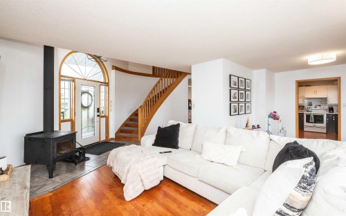 Hardwood flooring extending throughout the living space, a white entry door with an arched transom, a wood stove, a curved staircase with wood banister, and a kitchen visible through a doorway - 5502 60 Avenue, Barrhead, AB - Indoor