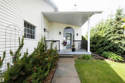 Inviting front entrance with a covered porch, arched transom window, white vinyl siding, black railings, and mature landscaping - 5502 60 Avenue, Barrhead, AB - Outdoor With Exterior