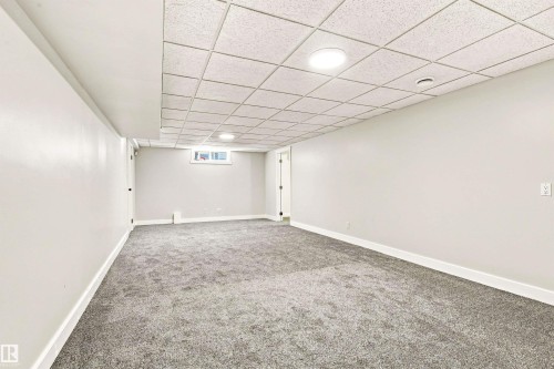Expansive room featuring light gray carpet, white walls, a drop ceiling with integrated lighting, and a white-framed window - 14307 50 Avenue Nw, Edmonton, AB 