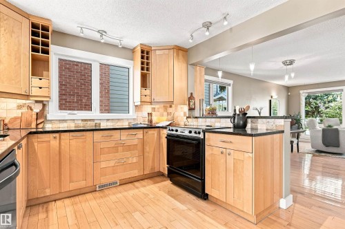 Kitchen featuring light wood cabinetry, black countertops, tile backsplash, stainless steel appliances, and wood-finish flooring - 14307 50 Avenue Nw, Edmonton, AB 