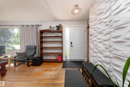 Welcoming entryway featuring hardwood flooring, textured 3D wall paneling, natural light from large windows with thermal-lined curtains, a white six-panel entry door, and built-in wooden shelving with open storage - 14906 76 Avenue Nw, Edmonton, AB 
