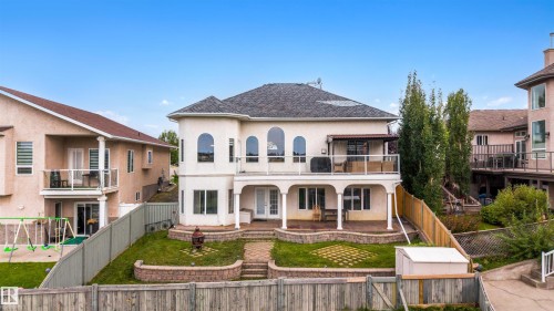 Multi-level exterior with stucco finish, upper-level balcony featuring glass railings, a covered patio with columns, lower-level patio with stone pavers, and a terraced backyard - 282 Ozerna Road Nw, Edmonton, AB 