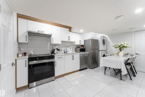 White cabinetry with wood-finish trim, stainless steel appliances, stacked laundry units, and white tile flooring - 13435 101 Street Nw, Edmonton, AB 