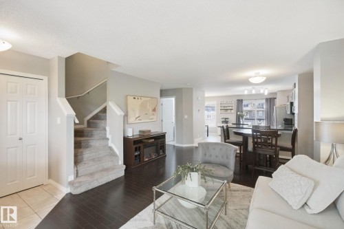 Dark wood-finish flooring, carpeted staircase with white railing, built-in media console, kitchen island with seating, and stainless steel appliances - #58 4029 Orchards Dr Sw, Edmonton, AB 