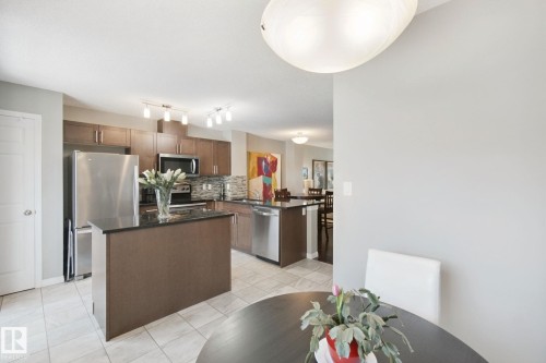 Kitchen featuring stainless steel appliances, dark wood-finish cabinetry, a central island with a dark countertop, and a tiled backsplash - #58 4029 Orchards Dr Sw, Edmonton, AB 