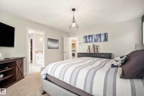 Carpeted bedroom featuring a dark wood-finish media console, a geometric pendant light fixture, and an entry to an additional room with a white vanity - 5966 Edmonds Crescent Nw, Edmonton, AB - Indoor Photo Showing Bedroom