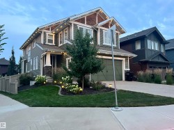 Two-story residence featuring green siding with white trim, a covered entry porch with stone accents, a two-car garage, and established landscaping with accent lighting - 
