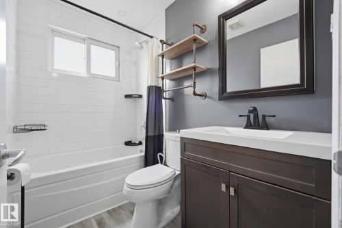 White subway tile shower surround, window with frosted glass, wood-finish flooring, dark wood vanity with integrated sink, and black-finish faucet - 13304 63 Street Nw, Edmonton, AB 